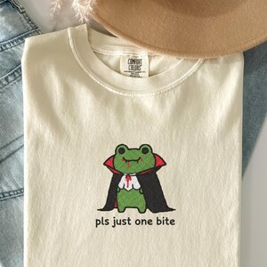 May include: A cream-coloured t-shirt with an embroidered green frog dressed as a vampire, wearing a black cape and red accents. The text "pls just one bite" is below the frog. A casual, Halloween-themed garment.