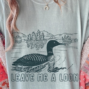 Peut inclure: T-shirt vert clair avec un dessin d'un huard nageant sur un lac, avec des montagnes en arrière-plan. Le texte "LEAVE ME A LOON" est imprimé en dessous. Le t-shirt est de style décontracté.