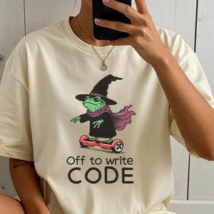 Op de afbeelding: Crèmekleurig T-shirt met een groene kikker met een heksenhoed, sjaal en zwarte jurk, op een rood hoverboard. De tekst "Off to write CODE" staat eronder.