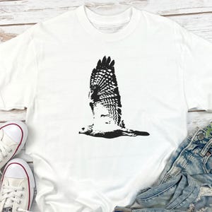 May include: White t-shirt featuring a black graphic of a hawk in flight. The bird's wings are spread wide, showcasing detailed feather patterns. The shirt is made of a soft material and is suitable for casual wear.