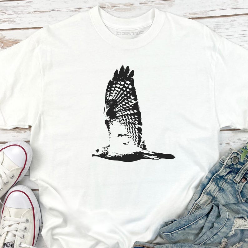 May include: White t-shirt featuring a black graphic of a hawk in flight. The bird's wings are spread wide, showcasing detailed feather patterns. The shirt is made of a soft material and is suitable for casual wear.