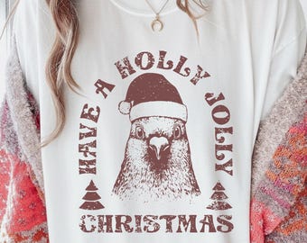 Könnte beinhalten: Weißes T-Shirt mit einem braunen Aufdruck eines Vogels mit Weihnachtsmütze. Der Text "HAVE A HOLLY JOLLY CHRISTMAS" ist in Braun um das Vogelbild herum gedruckt, mit zwei kleinen Weihnachtsbäumen unten.