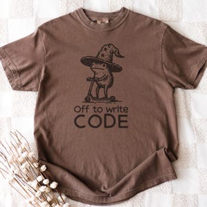 May include: Brown t-shirt featuring a whimsical illustration of a frog wearing a wizard hat and riding a scooter. The text "Off to write CODE" is printed below the graphic. The shirt is made of a soft material.
