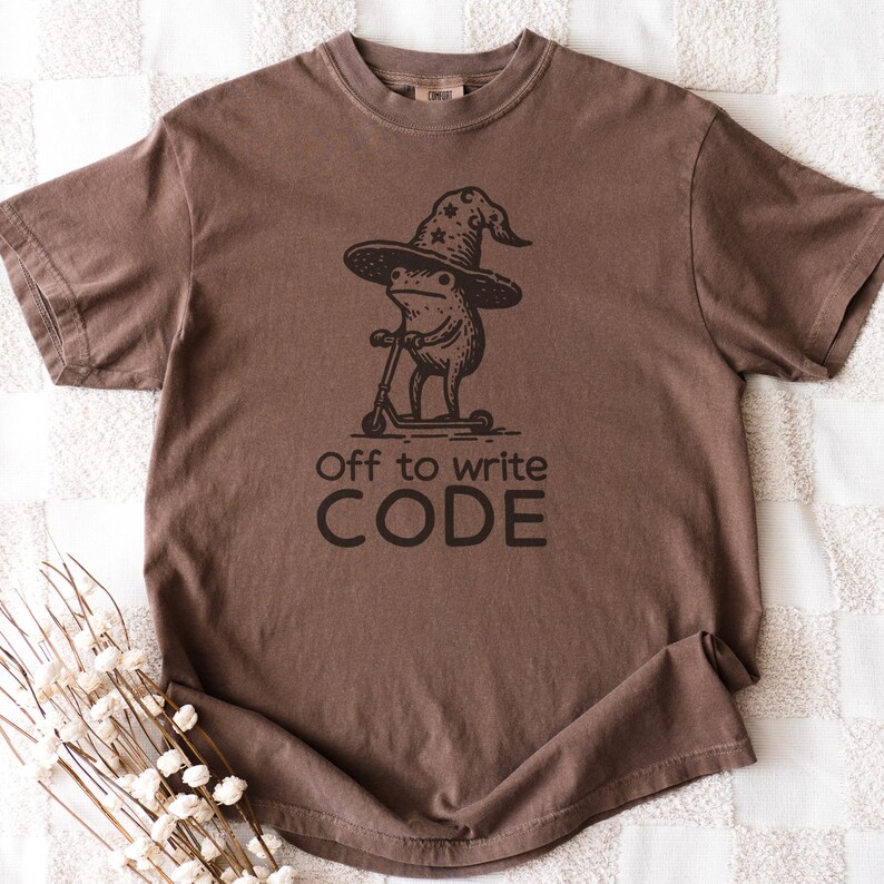 May include: Brown t-shirt featuring a whimsical illustration of a frog wearing a wizard hat and riding a scooter. The text "Off to write CODE" is printed below the graphic. The shirt is made of a soft material.