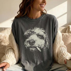 Può includere: T-shirt grigio scuro con un'illustrazione in bianco e nero di un cane con una sigaretta in bocca. Il volto del cane è dettagliato. La maglietta ha una vestibilità ampia.