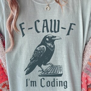 May include: A pale blue t-shirt featuring the text "F-CAW-F" above a black crow illustration. The crow is standing on a keyboard, with the text "I'm Coding" beneath. The shirt is made of a soft material.
