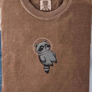 May include: A brown Comfort Colors t-shirt featuring an embroidered raccoon in a space suit design. The raccoon is gray and white, set within a white circle. The shirt is size XL-TG-EG.