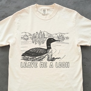 Peut inclure: T-shirt crème avec une illustration en noir et blanc d'un huard nageant dans un lac, avec des montagnes et des arbres en arrière-plan. Le texte "LEAVE ME A LOON" est imprimé en dessous.