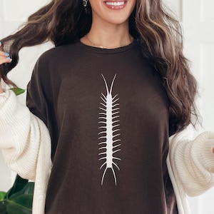 Puede incluir: Camiseta marrón oscuro con un diseño de ciempiés blanco. La camiseta está hecha de un material suave y forma parte de una colección de ropa. El diseño está centrado en la parte delantera.