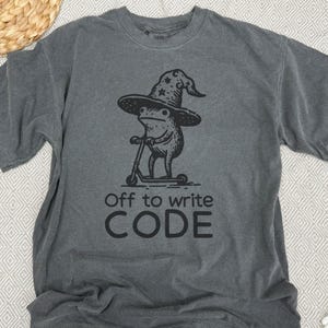 May include: Gray t-shirt featuring a whimsical illustration of a frog wearing a wizard hat and riding a scooter. The text "Off to write CODE" is printed below the graphic. The shirt is made of a soft material.