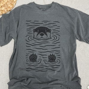 May include: Dark gray t-shirt featuring a black graphic of an otter in water. The otter's head and paws are visible, surrounded by wavy lines. The shirt has a crew neck and short sleeves, made of soft material.