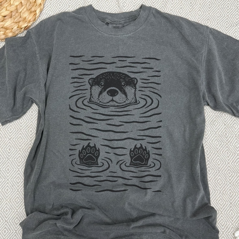 May include: Dark gray t-shirt featuring a black graphic of an otter in water. The otter's head and paws are visible, surrounded by wavy lines. The shirt has a crew neck and short sleeves, made of soft material.