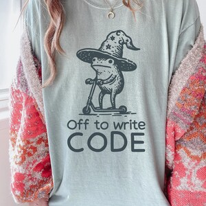 May include: A light blue t-shirt with a graphic of a frog wearing a wizard hat and riding a scooter. The text "Off to write CODE" is printed below the graphic. The shirt is a comfortable, casual style.