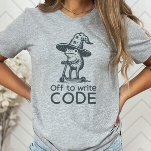 May include: A light gray t-shirt featuring a whimsical illustration of a frog wearing a wizard hat and riding a scooter. The text "Off to write CODE" is printed below the frog. The design is in dark gray.