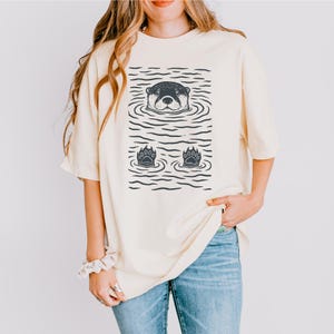 May include: Cream-colored t-shirt featuring a graphic of an otter in water. The otter's head and paws are visible, with water lines creating a textured effect. The shirt is a relaxed fit, suitable for casual wear.