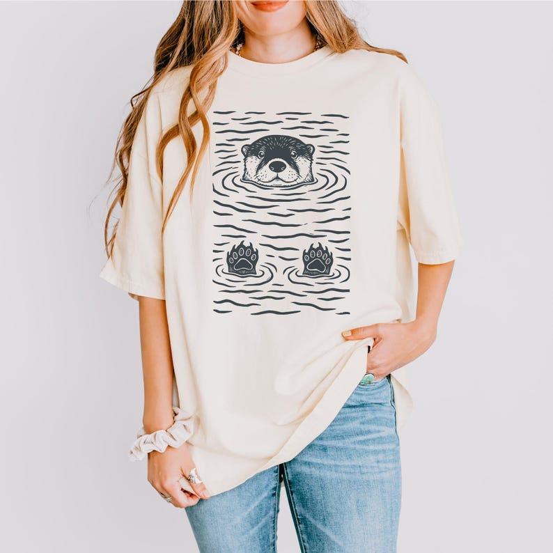 May include: Cream-colored t-shirt featuring a graphic of an otter in water. The otter's head and paws are visible, with water lines creating a textured effect. The shirt is a relaxed fit, suitable for casual wear.