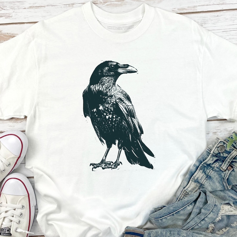 May include: White t-shirt featuring a detailed black illustration of a raven. The bird is depicted in a standing pose, with intricate feather detailing. The shirt has a classic crew neck design and is made of a soft, comfortable material.