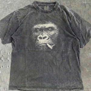 Puede incluir: Camiseta gris carbón con un gráfico detallado de un gorila fumando un cigarrillo. La cara del gorila está en escala de grises, con el cigarrillo en la boca. La camiseta tiene un aspecto vintage y lavado.