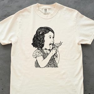Puede incluir: Camiseta de color crema con una ilustraci&oacute;n en blanco y negro de una ni&ntilde;a comiendo una babosa. El gr&aacute;fico est&aacute; centrado en la camiseta y representa a la ni&ntilde;a con la boca abierta, sosteniendo la babosa. La camiseta tiene cuello redondo y mangas cortas.