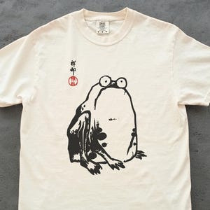 May include: Off-white t-shirt featuring a large black and white illustration of a frog. The frog has large eyes and is sitting. Above the frog is Japanese text and a red circle. The shirt is made of a soft material.