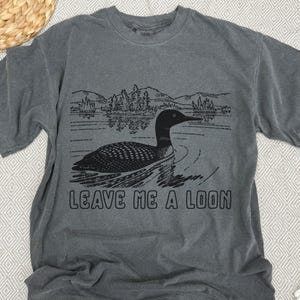 May include: Gray t-shirt with a graphic of a loon swimming on a lake, with trees and mountains in the background. The text "LEAVE ME A LOON" is printed below the image. The shirt has short sleeves and a crew neck.