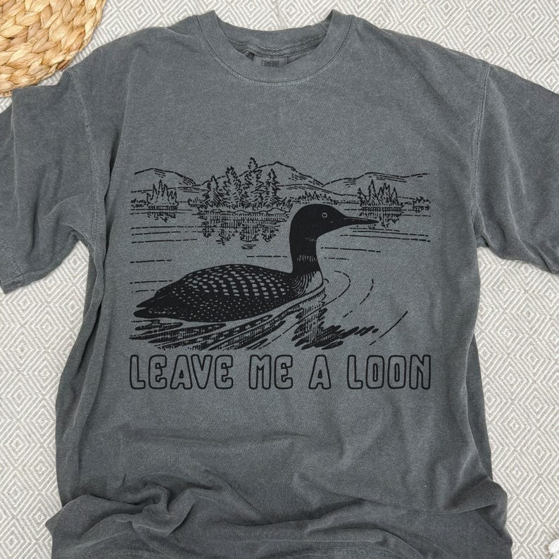 May include: Gray t-shirt with a graphic of a loon swimming on a lake, with trees and mountains in the background. The text "LEAVE ME A LOON" is printed below the image. The shirt has short sleeves and a crew neck.