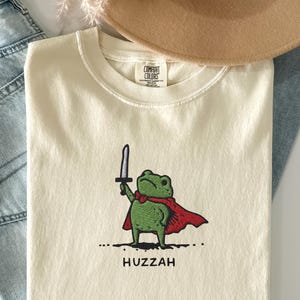 Può includere: T-shirt color crema con una rana verde con mantello rosso e spada. La parola "Huzzah" è stampata sotto. La maglietta è piegata, con un cappello beige e jeans blu sullo sfondo.