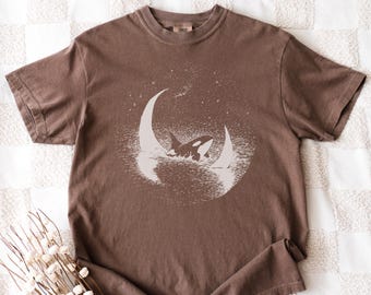 May include: A brown t-shirt with a white graphic of an orca whale swimming between two crescent moons. The design is set within a circle of stars and water. The shirt is made of a soft material.