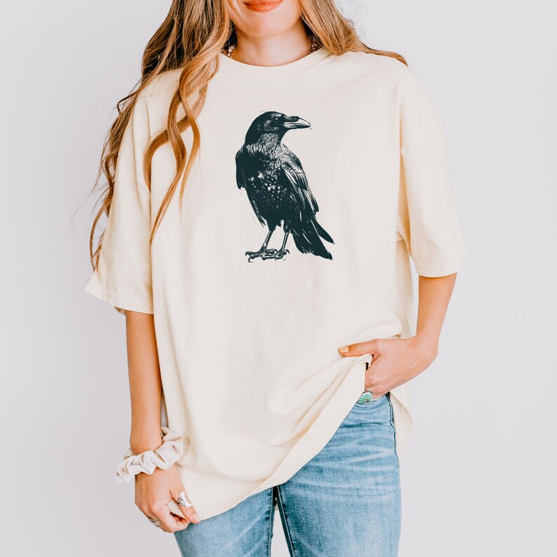 May include: Cream-colored t-shirt featuring a detailed black illustration of a raven. The bird is depicted in a standing pose, with its wings slightly folded. The shirt is a relaxed fit, with short sleeves.