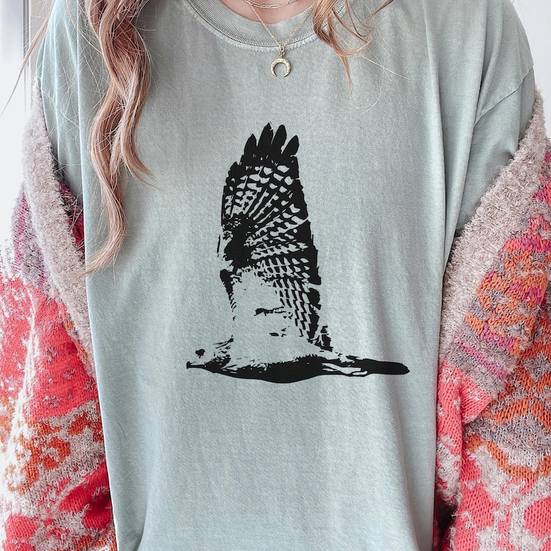May include: A light green t-shirt featuring a black graphic of a bird in flight. The bird's wings are spread wide, showcasing detailed feather patterns. The shirt is made of a soft material and has a relaxed fit, suitable for casual wear.
