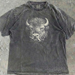 Può includere: T-shirt grigio antracite con una grafica dettagliata di un bisonte con una sigaretta. La grafica del bisonte è in una tonalità più chiara, creando contrasto con lo sfondo scuro della maglietta. Girocollo e maniche corte.