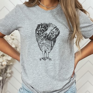 Puede incluir: Una camiseta gris claro con una ilustraci&oacute;n detallada en blanco y negro de un b&uacute;ho doblando la cabeza. Los ojos del b&uacute;ho son visibles y el dise&ntilde;o est&aacute; centrado en la camiseta.