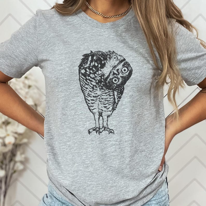 Puede incluir: Una camiseta gris claro con una ilustraci&oacute;n detallada en blanco y negro de un b&uacute;ho doblando la cabeza. Los ojos del b&uacute;ho son visibles y el dise&ntilde;o est&aacute; centrado en la camiseta.