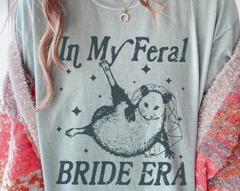 Pode incluir: Camiseta azul clara com o texto "In My Feral Bride Era" e um gráfico de um gambá usando um véu. O gambá está em uma pose relaxada com um braço levantado. O design inclui formas de diamante.