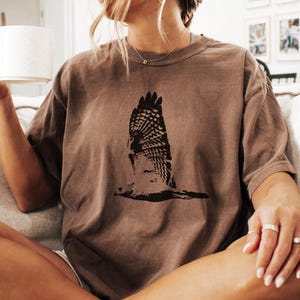 May include: A brown t-shirt featuring a black graphic of a bird in flight. The shirt has a relaxed fit and is made of a soft material. The design is centered on the front of the shirt, creating a striking visual.