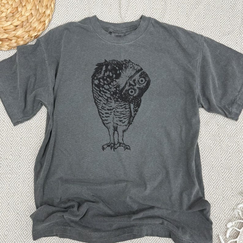 Puede incluir: Una camiseta gris con una ilustraci&oacute;n en blanco y negro de un b&uacute;ho mirando por encima del hombro. Las plumas del b&uacute;ho est&aacute;n detalladas, y la camiseta tiene un cuello redondo cl&aacute;sico y mangas cortas. La camiseta est&aacute; hecha de un material suave y c&oacute;modo.