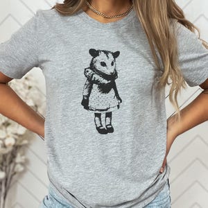 Pode incluir: Camiseta cinza com uma ilustra&ccedil;&atilde;o em preto e branco de um gamb&aacute; usando um vestido e botas. O gamb&aacute; &eacute; retratado em um estilo vintage, com um design detalhado. A camisa &eacute; feita de um material macio.