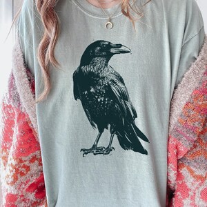 May include: A light green t-shirt featuring a detailed black illustration of a raven. The raven is depicted in a side profile, showcasing its beak and feathers. The shirt is a casual style.