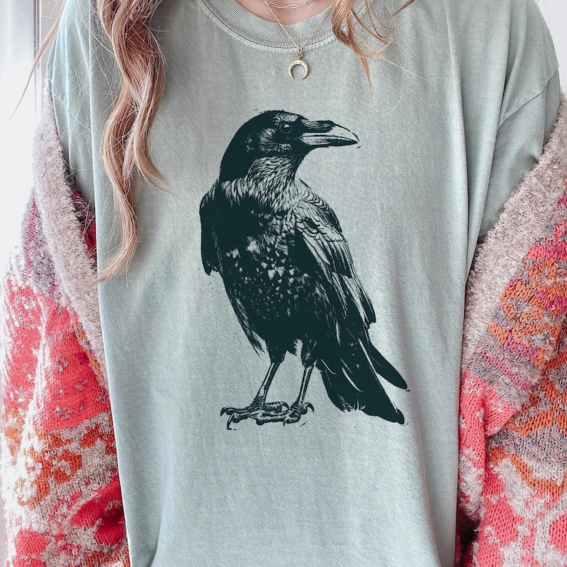 May include: A light green t-shirt featuring a detailed black illustration of a raven. The raven is depicted in a side profile, showcasing its beak and feathers. The shirt is a casual style.