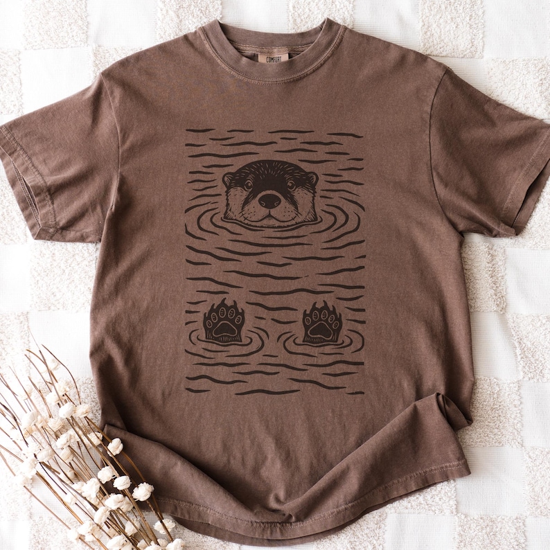 May include: Brown t-shirt featuring a black and brown graphic of an otter in water. The otter's head and paws are visible, with water lines surrounding them. The shirt has a crew neck and short sleeves.
