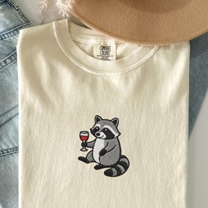 May include: A cream-coloured t-shirt with an embroidered raccoon holding a glass of red wine. The raccoon is in grey and black, with a striped tail. The shirt is folded, with a tan hat and blue jeans in the background.