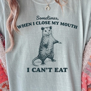 May include: A light green t-shirt with a graphic of an opossum and the text "Sometimes, when I close my mouth, I can't eat." The opossum is standing upright with its arms out. The shirt is a relaxed fit.