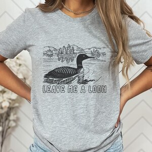 May include: A gray t-shirt featuring a black and white graphic of a loon swimming on a lake with mountains in the background. The text "LEAVE ME A LOON" is printed below the graphic.