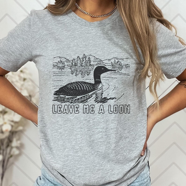 May include: A gray t-shirt featuring a black and white graphic of a loon swimming on a lake with mountains in the background. The text "LEAVE ME A LOON" is printed below the graphic.