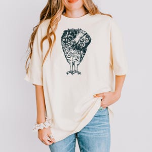 Puede incluir: Camiseta color crema con una ilustraci&oacute;n detallada en blanco y negro de un b&uacute;ho. El b&uacute;ho est&aacute; representado con la cabeza girada, mostrando sus ojos y plumas. La camiseta tiene un corte holgado y mangas cortas.