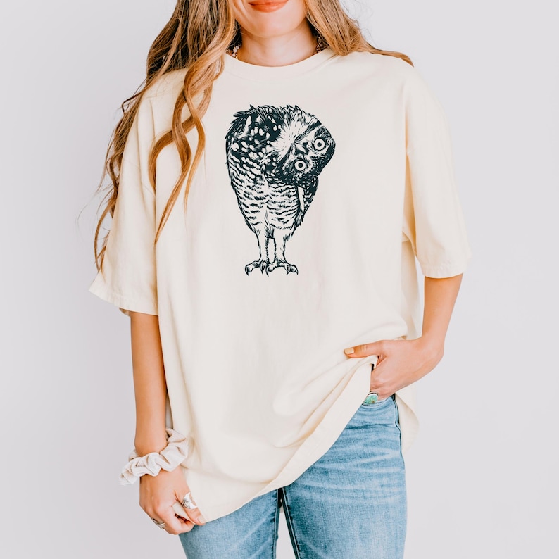 Puede incluir: Camiseta color crema con una ilustraci&oacute;n detallada en blanco y negro de un b&uacute;ho. El b&uacute;ho est&aacute; representado con la cabeza girada, mostrando sus ojos y plumas. La camiseta tiene un corte holgado y mangas cortas.