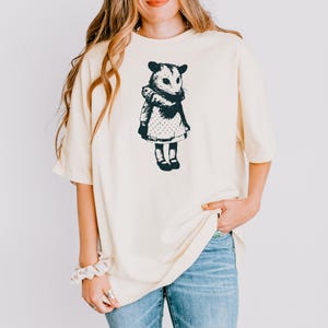 Pode incluir: Camiseta creme com uma ilustra&ccedil;&atilde;o em preto e branco de um gamb&aacute; usando um vestido. O gamb&aacute; &eacute; retratado em um estilo vintage, com um vestido e sapatos detalhados. A camisa tem um ajuste folgado e mangas curtas.
