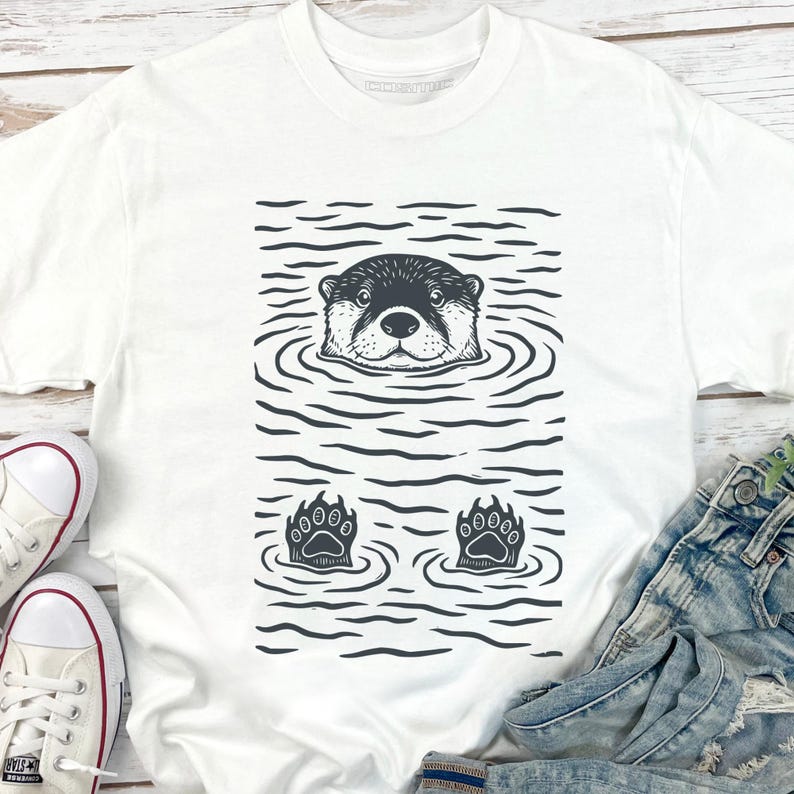 May include: White t-shirt featuring a black and white illustration of an otter in water. The otter's head and paws are visible, surrounded by water ripples. The design is centered on the shirt.