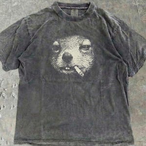 May include: A dark grey t-shirt with a white graphic of a squirrel smoking a cigarette. The t-shirt has a crew neck and short sleeves. The fabric has a faded, worn appearance.