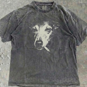 Puede incluir: Camiseta gris antracita con un estampado gráfico de un perro fumando un cigarrillo. La cara del perro es blanca sobre la tela gris oscura. La camiseta tiene cuello redondo y mangas cortas. La tela tiene un aspecto envejecido.